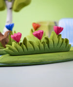 Bumbu large wooden grass with three pink flowers toy scenery piece placed at the front of a small world play scene with other flowers and trees in the background.
