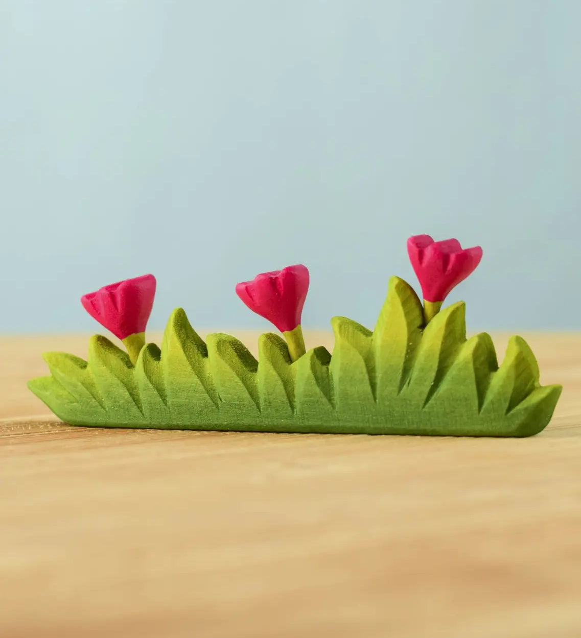 Bumbu large wooden grass with three pink flowers toy scenery piece placed on a wooden surface. 