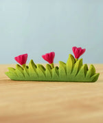 Bumbu large wooden grass with three pink flowers toy scenery piece placed on a wooden surface. 