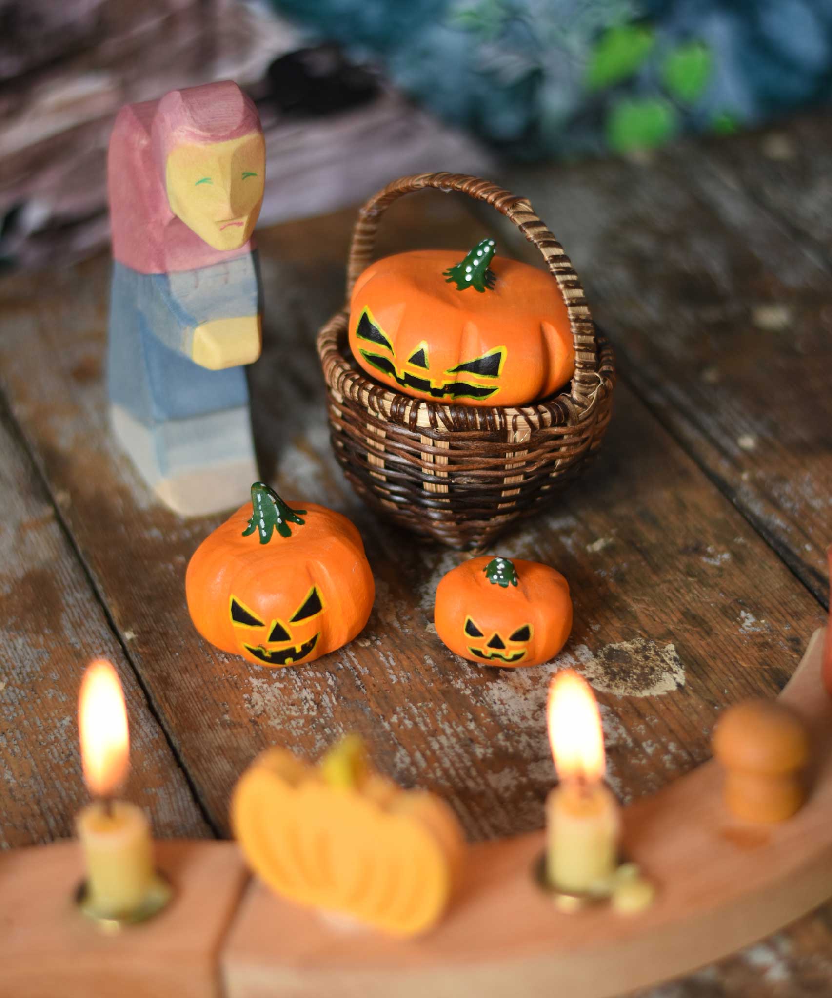 Bumbu Halloween pumpkins placed in and around a Kraul basket on a wooden surface. There is a Ostheimer wooden witch figure besides them and a decorated Grimm's celebration ring with lit candle in front of them. These wooden figures are part of a wide range of expertly hand crafted wooden toys here at Babipur.