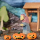 A collection of three Bumbu Halloween pumpkins placed in a line on a wooden surface at the front of a small world play scene. An Ostheimer witch can be seen in the background along with a wooden water well and birch tree. These wooden figures are part of a wide range of expertly hand crafted wooden toys here at Babipur. 
