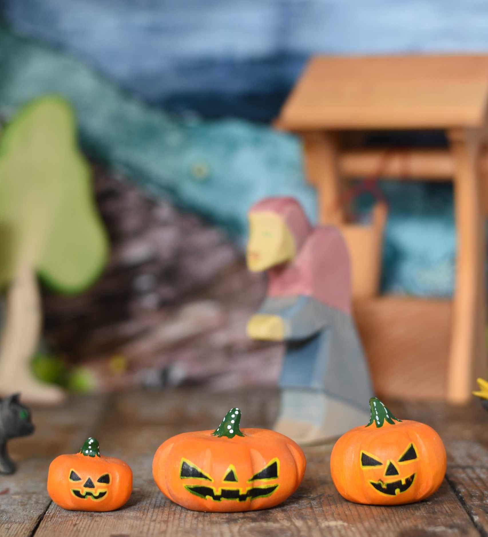 A collection of three Bumbu Halloween pumpkins placed in a line on a wooden surface at the front of a small world play scene. An Ostheimer witch can be seen in the background along with a wooden water well and birch tree. These wooden figures are part of a wide range of expertly hand crafted wooden toys here at Babipur. 
