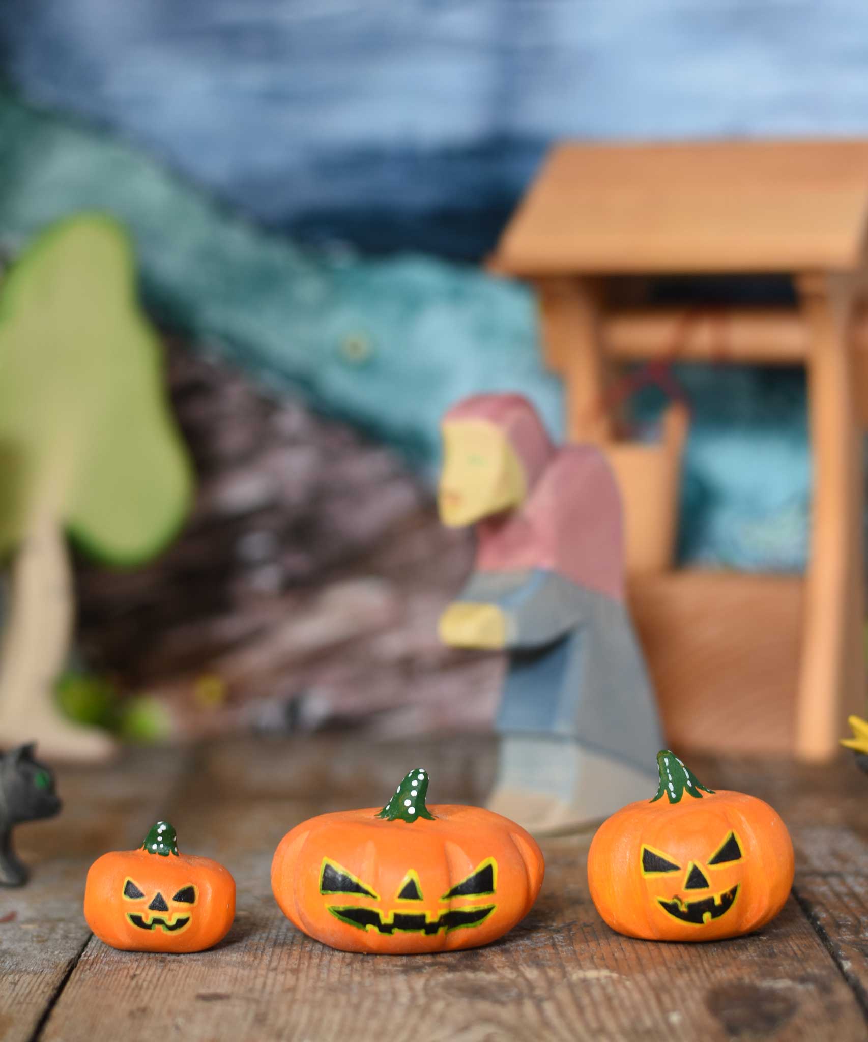 A collection of three Bumbu Halloween pumpkins placed in a line on a wooden surface at the front of a small world play scene. An Ostheimer witch can be seen in the background along with a wooden water well and birch tree. These wooden figures are part of a wide range of expertly hand crafted wooden toys here at Babipur. 

