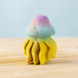 The Bumbu hand carved wooden jellyfish toy. It is has a rounded bell-shape head which is hand painted in ombre pink and blue, with bright yellow tentacles. 