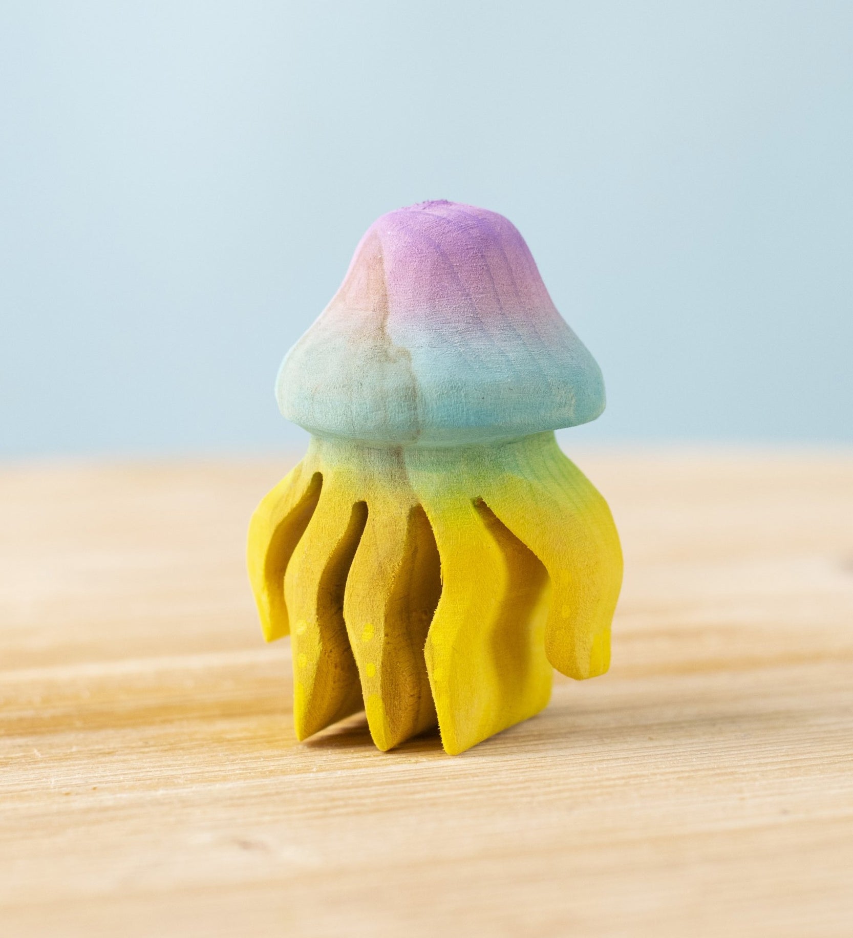 The Bumbu hand carved wooden jellyfish toy. It is has a rounded bell-shape head which is hand painted in ombre pink and blue, with bright yellow tentacles. 