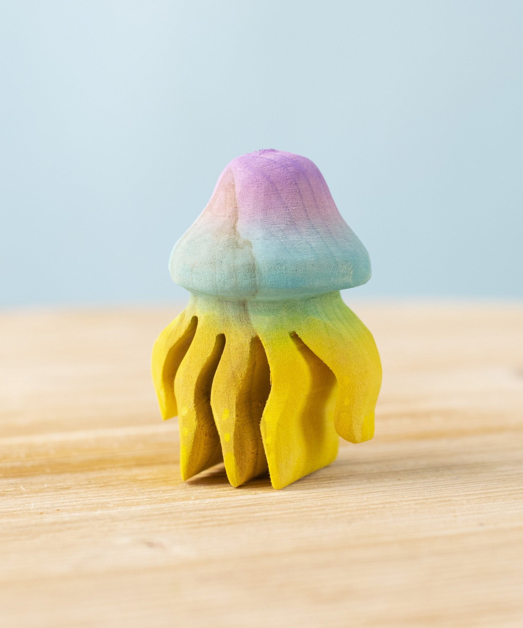 The Bumbu hand carved wooden jellyfish toy. It is has a rounded bell-shape head which is hand painted in ombre pink and blue, with bright yellow tentacles. 