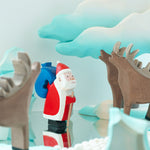Bumbu children's wooden Father Christmas figure stood on a mirror next to some Bumbu stags and a winter Maple tree