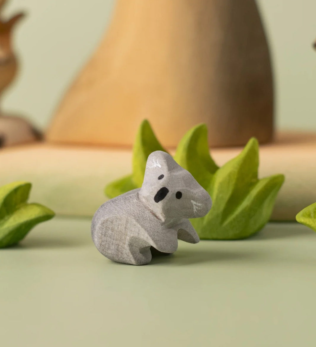 A Bumbu Toys handmade wooden Koala Cub figure placed on a green coloured surface with wooden scenery and greenery pieces in the background. The adult koala figure and scenery pieces are available at Babipur. 