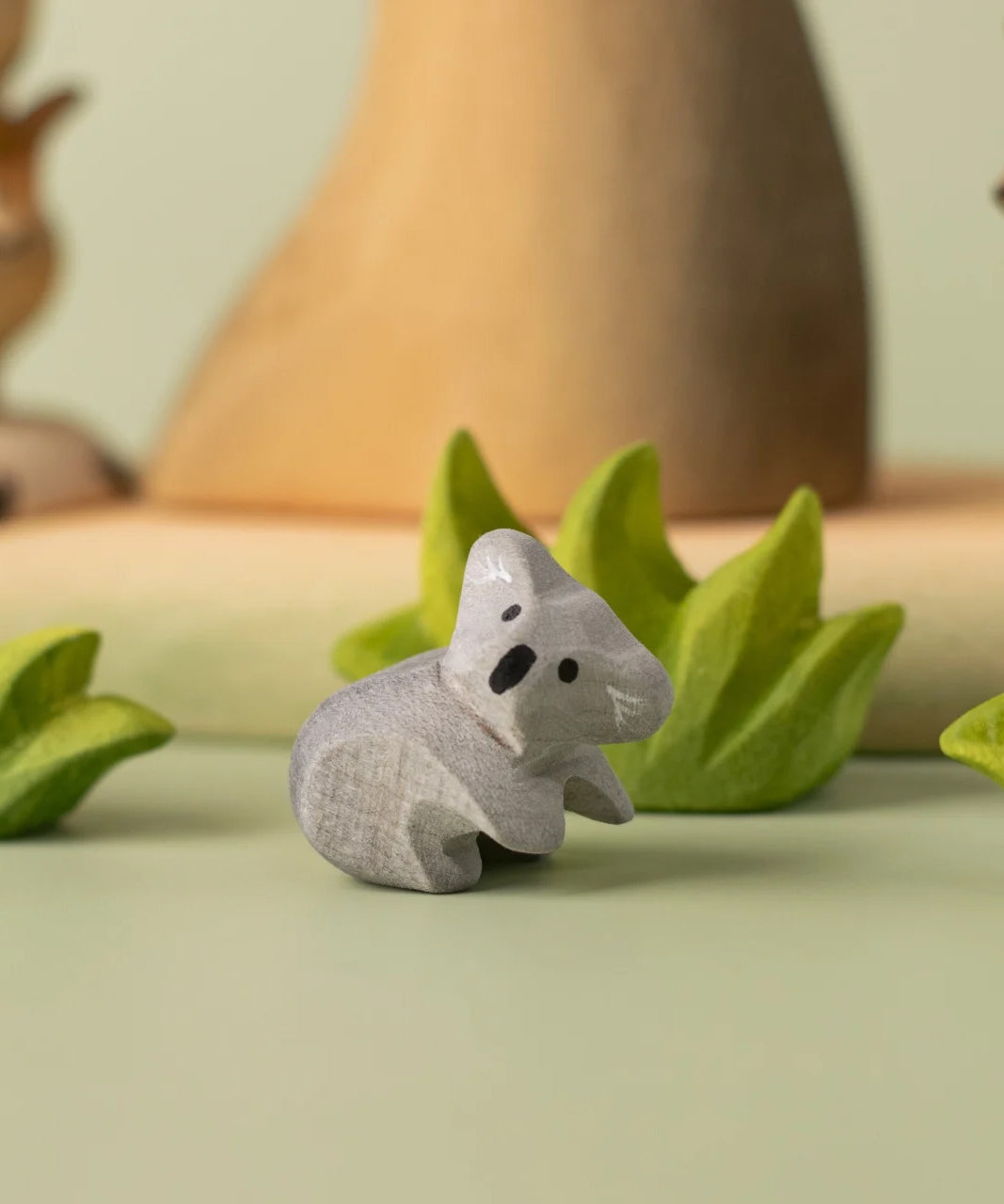 A Bumbu Toys handmade wooden Koala Cub figure placed on a green coloured surface with wooden scenery and greenery pieces in the background. The adult koala figure and scenery pieces are available at Babipur. 