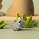 A Bumbu Toys handmade wooden Koala Cub figure placed on a green coloured surface with wooden scenery and greenery pieces in the background. The adult koala figure and scenery pieces are available at Babipur. 