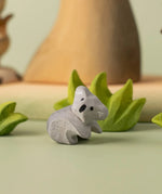 A Bumbu Toys handmade wooden Koala Cub figure placed on a green coloured surface with wooden scenery and greenery pieces in the background. The adult koala figure and scenery pieces are available at Babipur. 