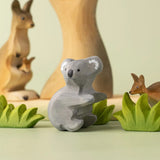 A Bumbu Toys handmade wooden Koala figure placed on a green surface surrounded by wooden greenery pieces and a eucalyptus tree and kangaroos in the background. 