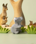 A Bumbu Toys handmade wooden Koala figure placed on a green surface surrounded by wooden greenery pieces and a eucalyptus tree and kangaroos in the background. 