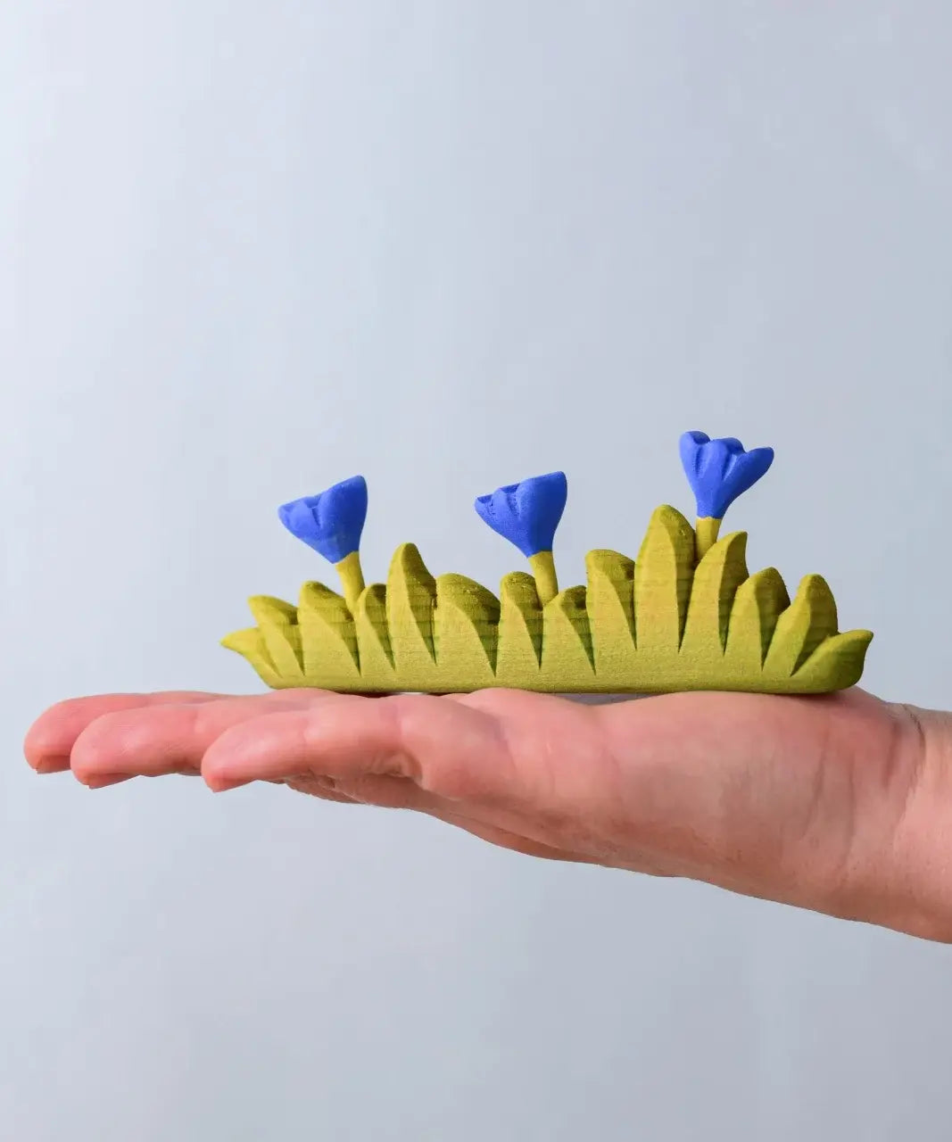 Bumbu large grass with 3 blue flowers wooden toy scenery piece in an adult's hand. 