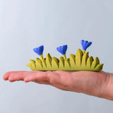 Bumbu large grass with 3 blue flowers wooden toy scenery piece in an adult's hand. 