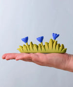 Bumbu large grass with 3 blue flowers wooden toy scenery piece in an adult's hand. 