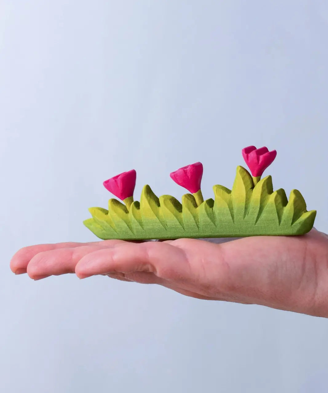 Bumbu large wooden grass with three pink flowers toy scenery piece in an adult's hand. 