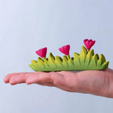  Bumbu large wooden grass with three pink flowers toy scenery piece in an adult's hand. 