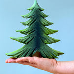 A large Bumbu wooden Fir tree balanced on an adult's hand against a blue background.