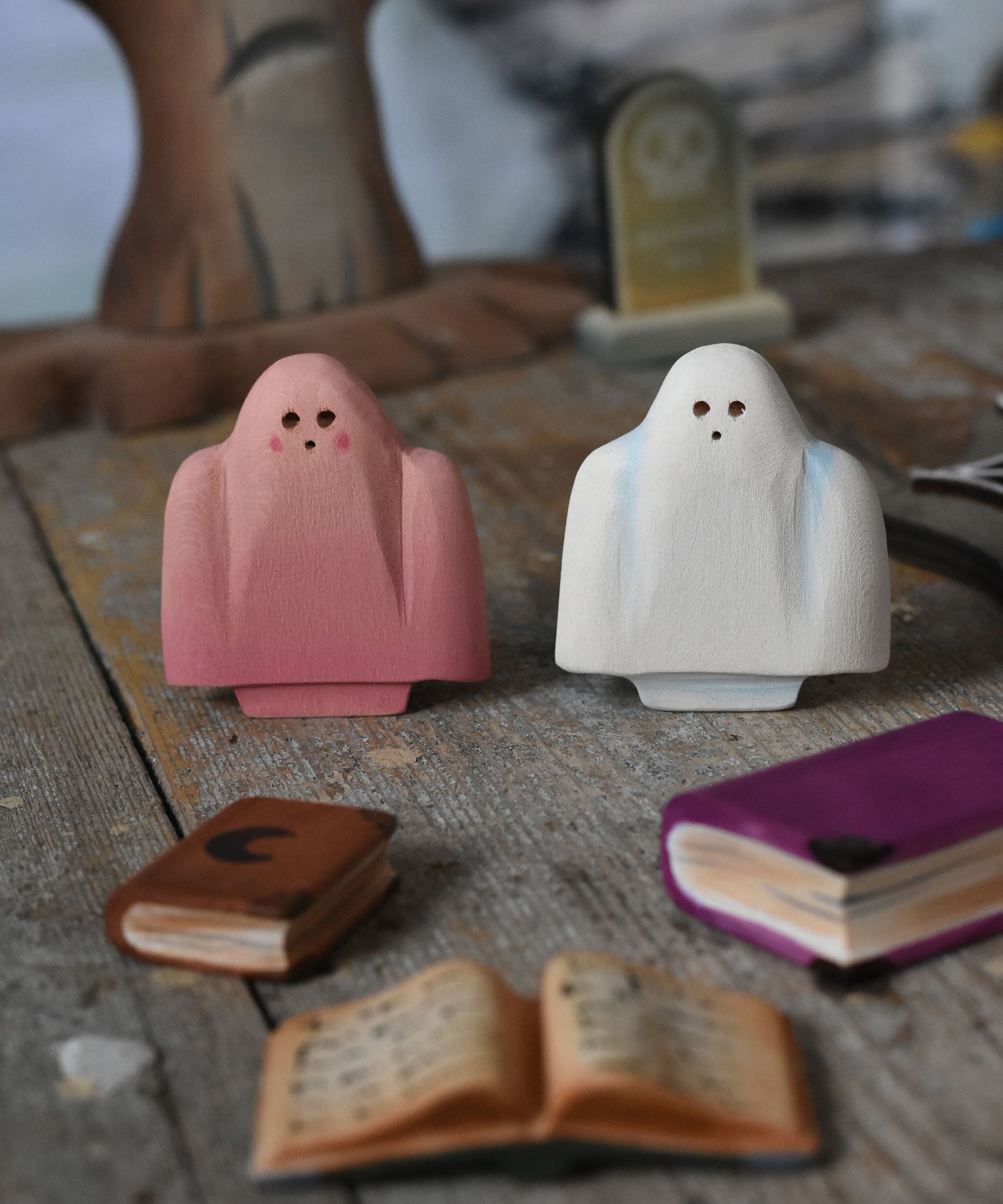 A pink and white Bumbu ghost placed on a wooden surface in a small world play scene. There are little wooden books in front of them and a spooky tree and a gravestone in the background. These expertly hand crafted items are part of a large range of wooden toys available here at Babipur. 