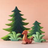 A Bumbu Toys Baby Dragon wooden figure placed at the front of a small world play scene. There are Bumbu wooden trees and greenery pieces in the background. 