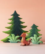 A Bumbu Toys Baby Dragon wooden figure placed at the front of a small world play scene. There are Bumbu wooden trees and greenery pieces in the background. 