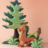 A Bumbu dragon toy placed in the middle of a small world play scene with a Bumbu tree and greenery. These are part of a large collection of handmade wooden toy animals and scenery pieces that are available here at Babipur. 