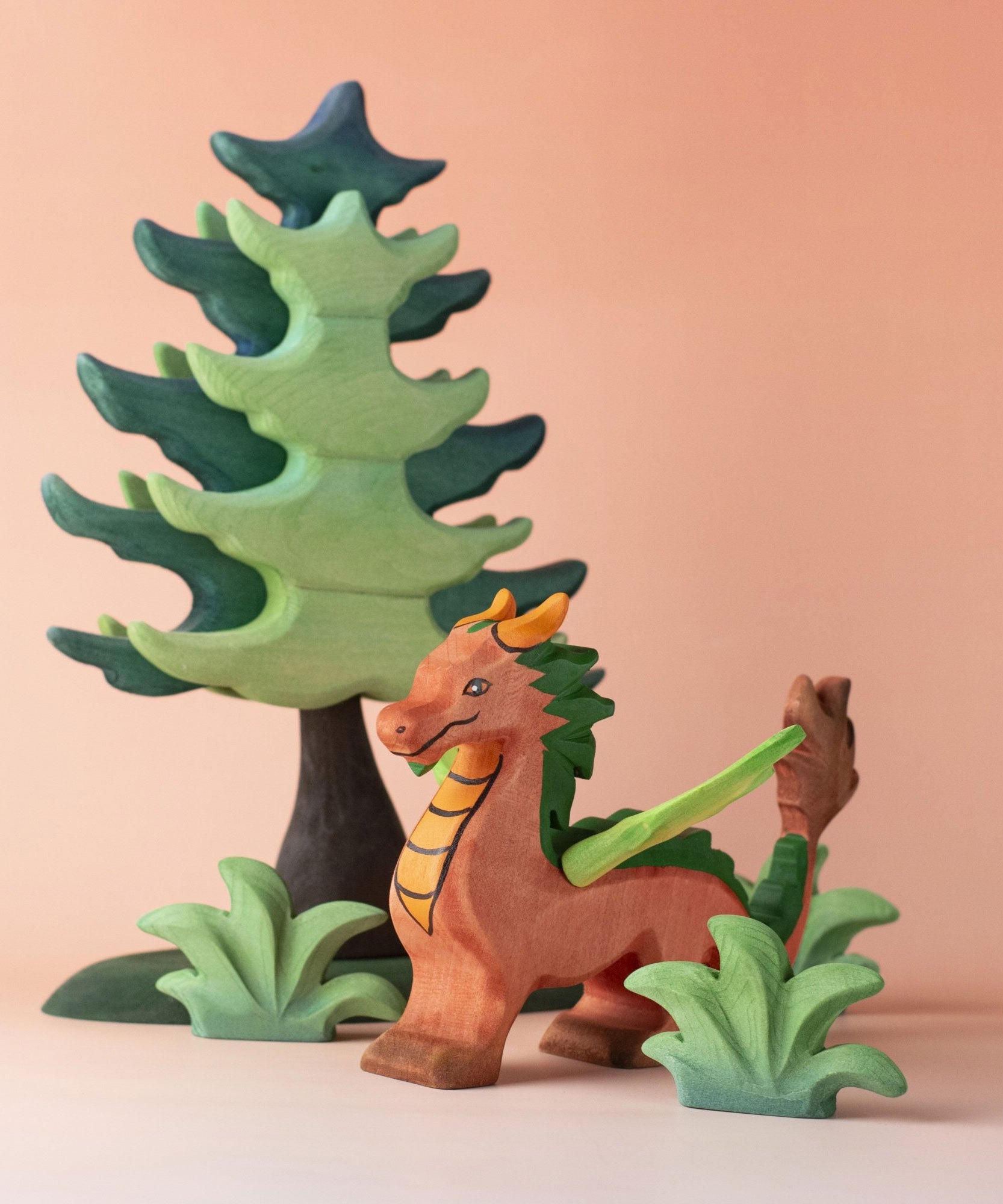 A Bumbu dragon toy placed in the middle of a small world play scene with a Bumbu tree and greenery. These are part of a large collection of handmade wooden toy animals and scenery pieces that are available here at Babipur. 
