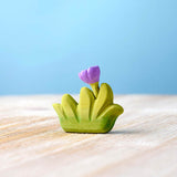 Bumbu handmade small wooden grass toy with lilac flower on a wooden worktop in front of a blue background