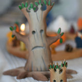A small Bumbu spooky tree figure placed on a light grey tablecloth, a large tree and celebration ring can be seen blurred out in the background.
