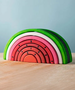 Bumbu Toys wooden stacking Watermelon toy placed on a wooden surface, showing the curved layered slices in vibrant pink, red and green. Part of the handmade wooden toy range from Bumbu available here at Babipur.
