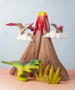 A small world play scene featuring handmade wooden toys from a brand called Bumbu. There are Pterandons and a t-rex standing on and around a wooden volcano and green coloured wooden foliage. 
