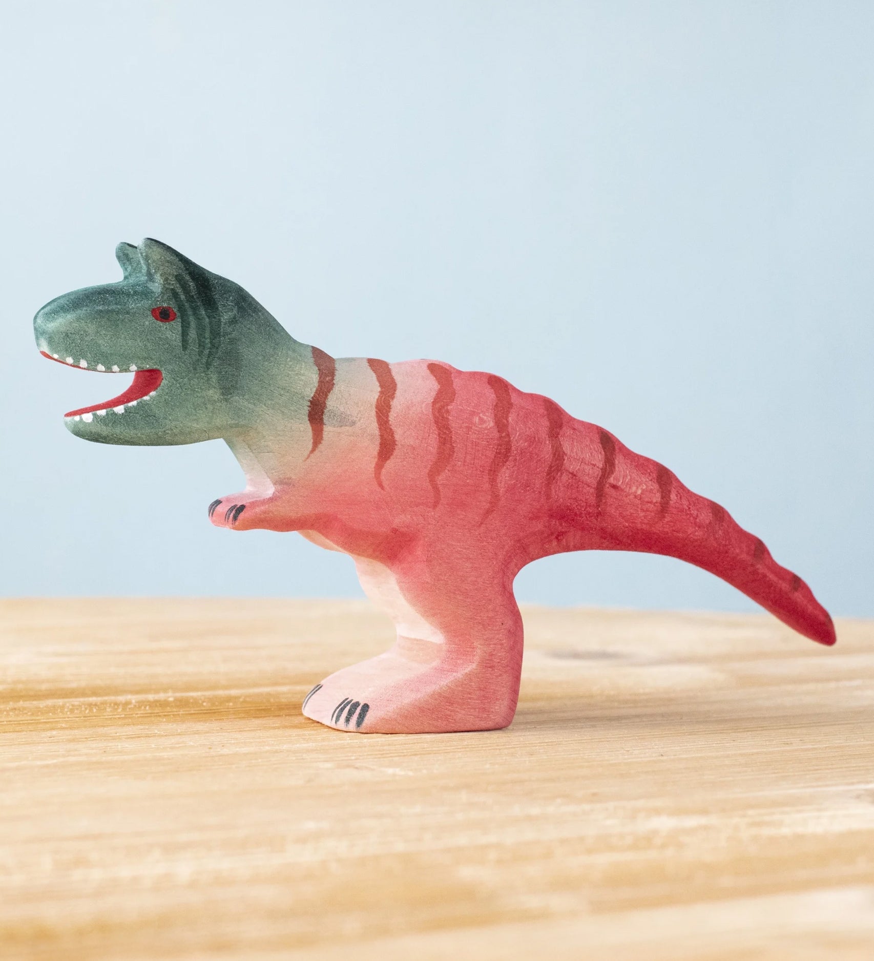 Bumbu Toys Big Carnotaurus dinosaur wooden figure placed on a wooden surface. The handmade dinosaur figure is part of a wide range of wooden animal figures here at Babipur. 