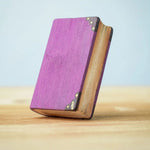 Bumbu Toys Large purple Book from their Halloween collection. The edge of the book can be seen showing yellowed pages which gives a spooky feel. The cover is a moody purple with black corner plates and gold details. 