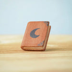 Bumbu Toys Small Book decorated with a black moon on the front. The book has dark brown corner plates and black dots. The book is propped up on a wooden surface.