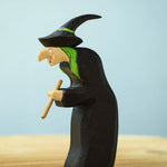 Bumbu Toys Brewing Witch wooden figure placed on a wooden surface. The witch has bright green hair and is wearing a black hat decorated with a painted bright green ribbon around the brim. The witch is clutching a wooden brewing stick in her hands. 