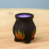 A Bumbu Wooden Witch's Cauldron. The cauldron is black on the outside and decorated with red, yellow and orange painted flames on the side. The top of the cauldron has a purple swirled effect decoration. The cauldron is placed on a wooden surface.