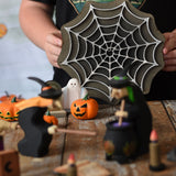 A child holding up the Bumbu wooden cobweb toy in the back of a small world play scene set up on a wooden surface. There are wooden witches, ghost and pumpkin figures in the scene. These expertly hand crafted items are part of a large range of wooden toys available here at Babipur. 