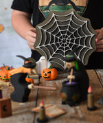 A child holding up the Bumbu wooden cobweb toy in the back of a small world play scene set up on a wooden surface. There are wooden witches, ghost and pumpkin figures in the scene. These expertly hand crafted items are part of a large range of wooden toys available here at Babipur. 