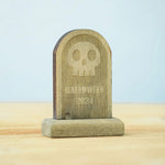 The skull decorated headstone from the Bumbu Toys Gravestones Set. The headstone is placed on a wooden surface.