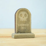 The skull decorated headstone from the Bumbu Toys Gravestones Set. The headstone is placed on a wooden surface.