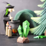 The green coloured Bumbu crystals wooden toy figure placed at the front of a small world play scene. Other Bumbu toys including a tree and witch figure can be seen blurred in the background. 