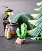 The green coloured Bumbu crystals wooden toy figure placed at the front of a small world play scene. Other Bumbu toys including a tree and witch figure can be seen blurred in the background. 