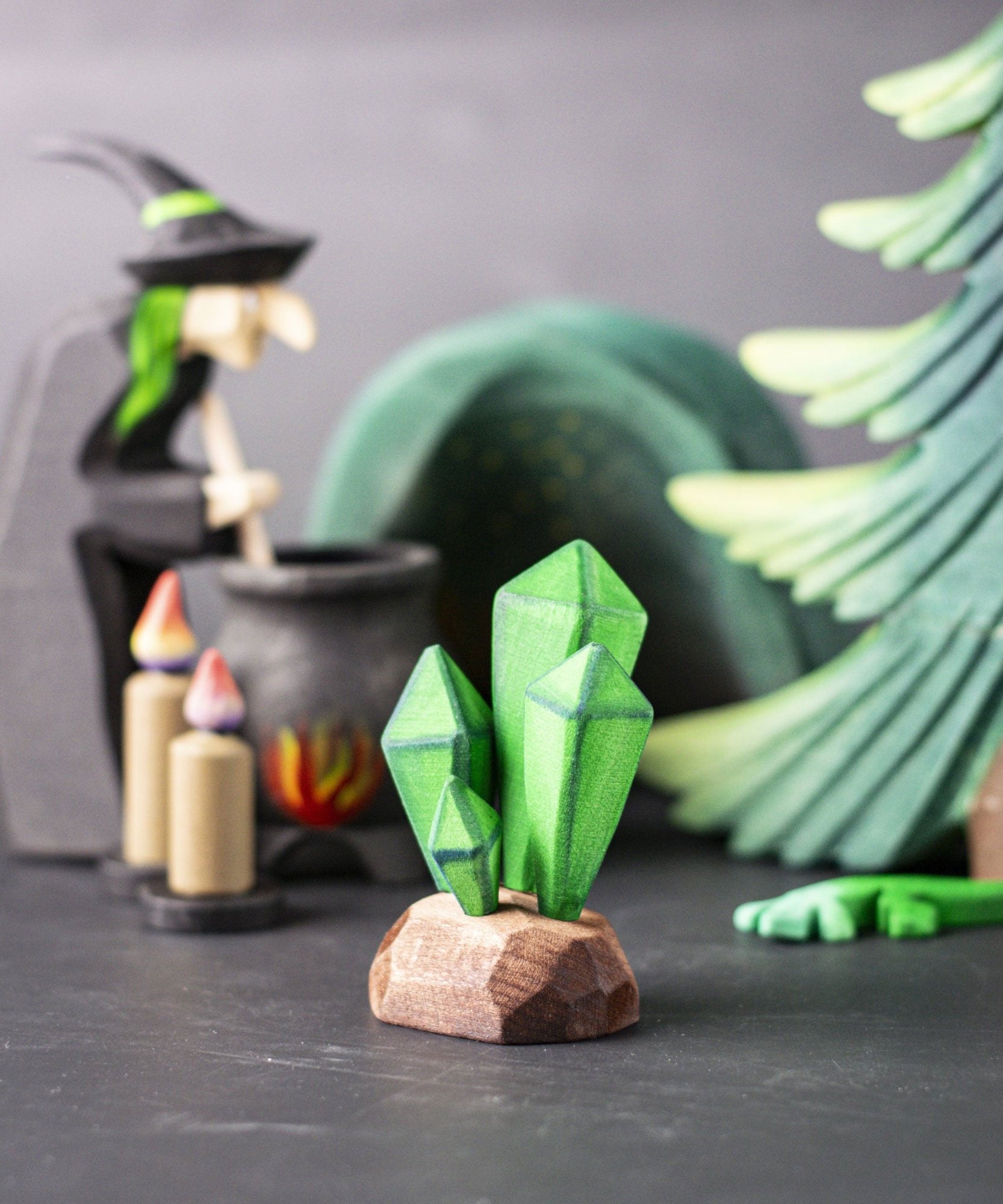 The green coloured Bumbu crystals wooden toy figure placed at the front of a small world play scene. Other Bumbu toys including a tree and witch figure can be seen blurred in the background. 