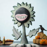 Bumbu Toys black wooden sunflower figure with a mouth with light pink lips and white teeth carved into it for a creepy Halloween vibe. The toy sunflower figure is placed on a wooden surface and surrounded by other Halloween themed wooden toys. 