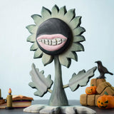 Bumbu Toys black wooden sunflower figure with a mouth with light pink lips and white teeth carved into it for a creepy Halloween vibe. The toy sunflower figure is placed on a wooden surface and surrounded by other Halloween themed wooden toys. 