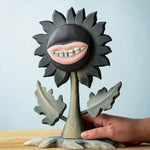 A hand reaching for the Bumbu Toys black wooden sunflower figure with a mouth with light pink lips and white teeth carved into it for a creepy Halloween vibe. The toy sunflower figure is placed on a wooden surface. 