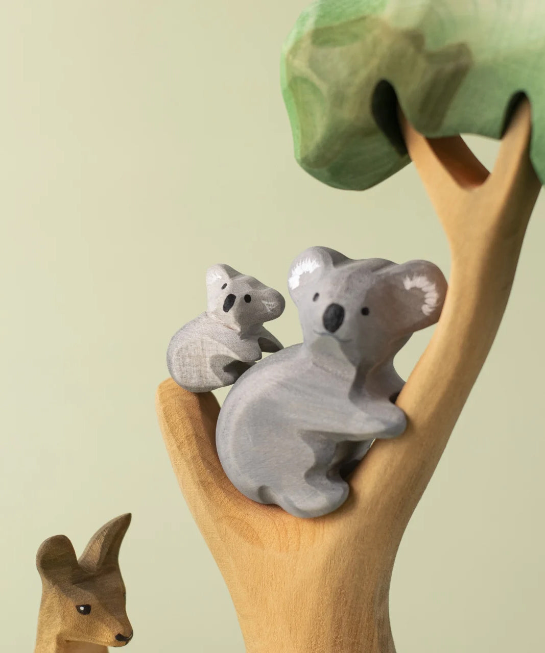 A Bumbu Toys handmade wooden Koala Cub figure and adult Koala placed on a branch of a wooden eucalyptus tree. The full Koala collection is available at Babipur.