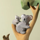 A Bumbu Toys handmade wooden Koala Cub figure and adult Koala placed on a branch of a wooden eucalyptus tree. The full Koala collection is available at Babipur.