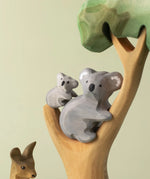 A Bumbu Toys handmade wooden Koala Cub figure and adult Koala placed on a branch of a wooden eucalyptus tree. The full Koala collection is available at Babipur.
